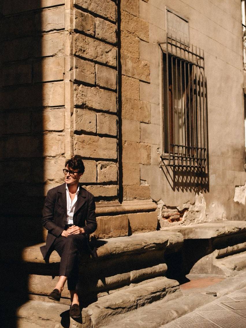 A man dressed in a stylish suit, sitting on stone steps with a historic building backdrop, enjoying the Pitti Uomo summer fashion event in Florence, Italy