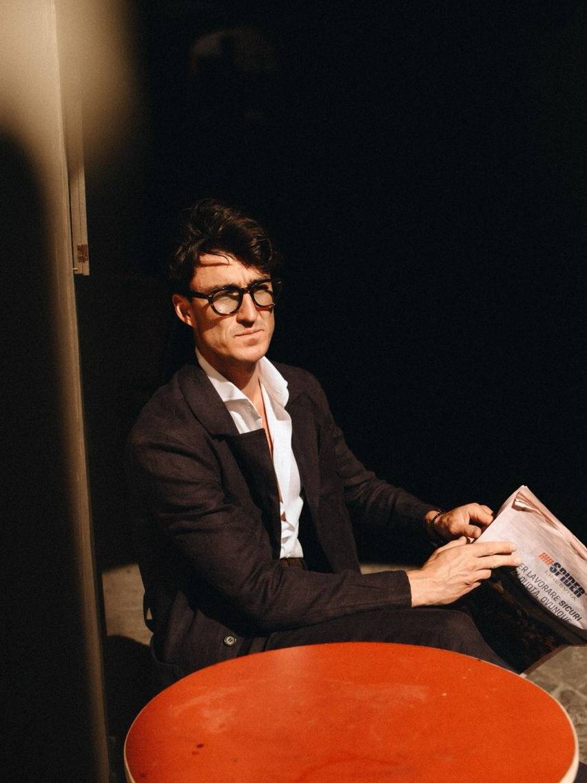 A man in a smart outfit sitting at a red table, reading a newspaper, epitomizing the style at the Pitti Uomo summer fashion event in Florence, Italy