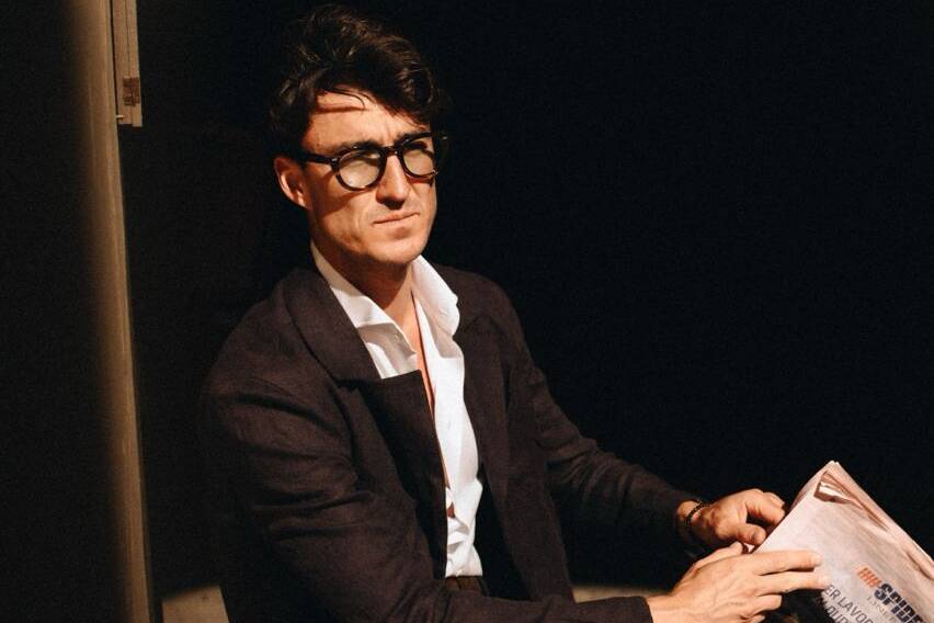 A man in a smart outfit sitting at a red table, reading a newspaper, epitomizing the style at the Pitti Uomo summer fashion event in Florence, Italy