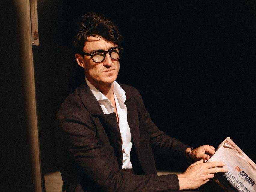 A man in a smart outfit sitting at a red table, reading a newspaper, epitomizing the style at the Pitti Uomo summer fashion event in Florence, Italy