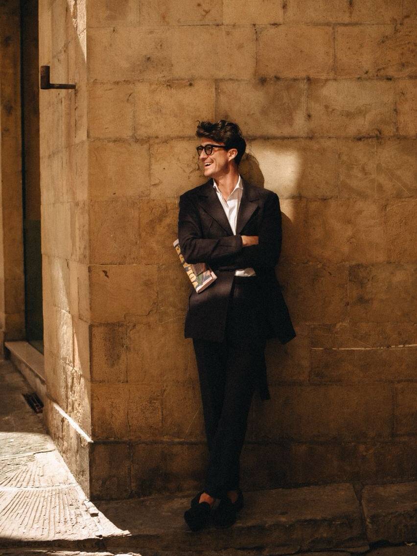 A man leaning against a rustic wall, smiling and holding a newspaper, capturing the essence of the Pitti Uomo summer fashion event in Florence, Italy