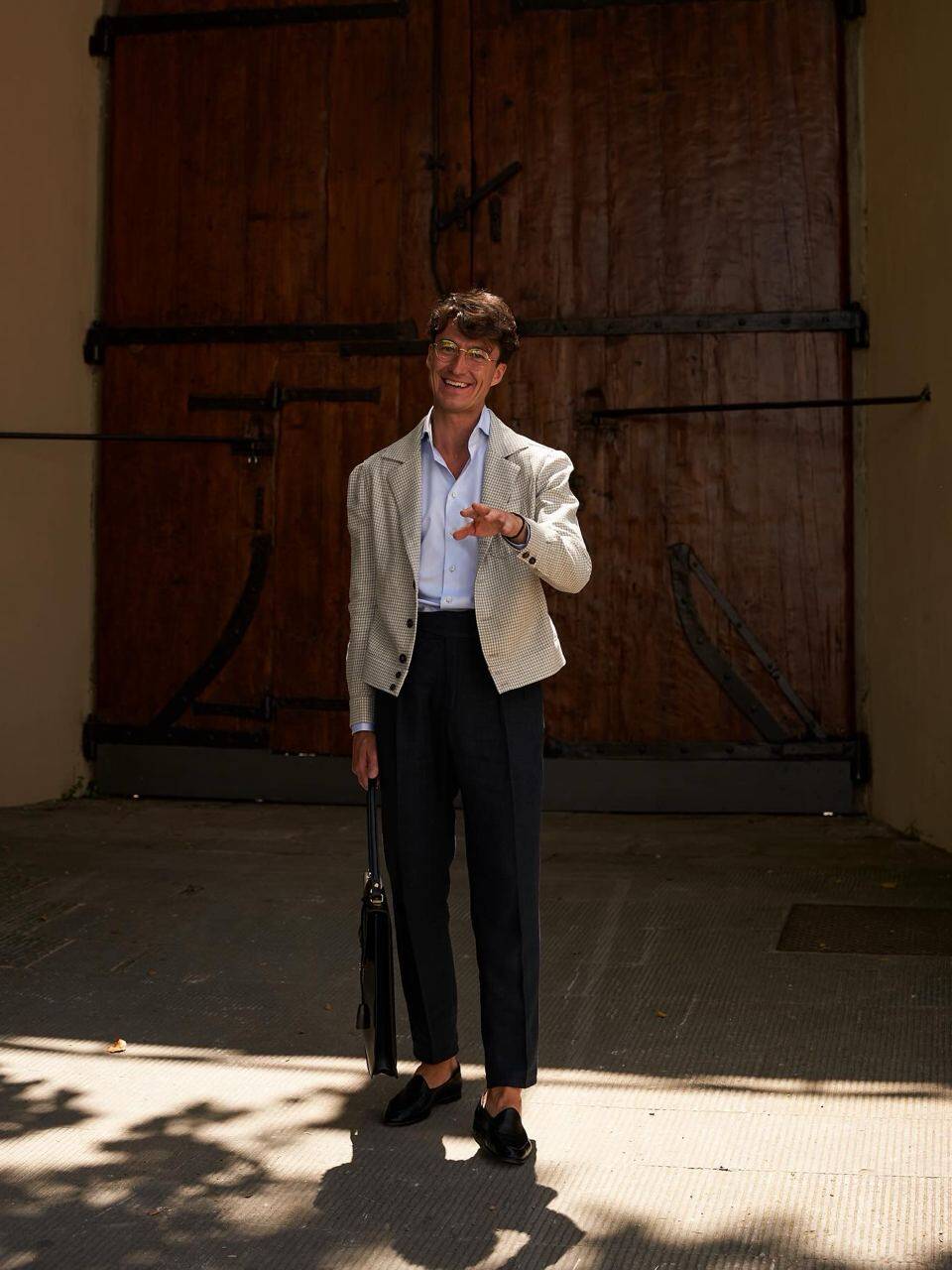 Anton Masko posing in front of large wooden doors, showcasing elegant attire with a leather briefcase