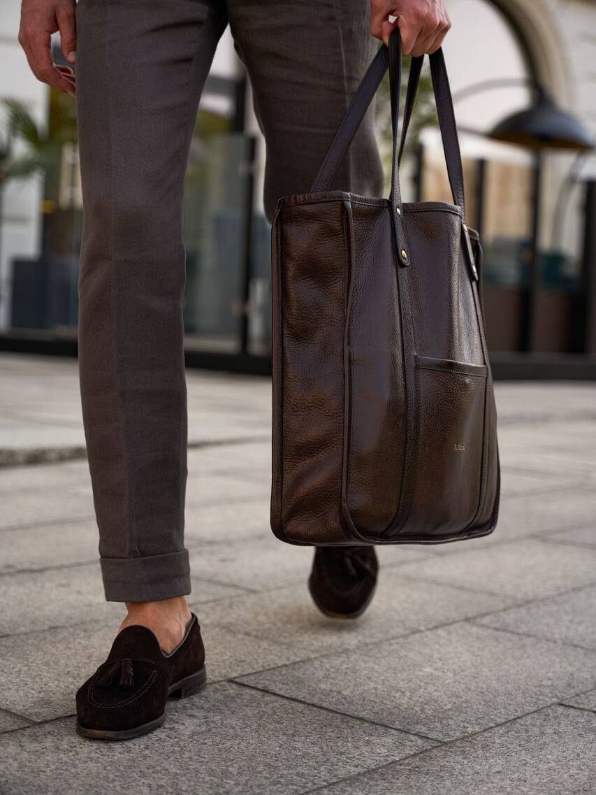 Close-up of a man carrying a handcrafted leather tote bag, showcasing leather craftsmanship. Brown leather bag with tassel loafers