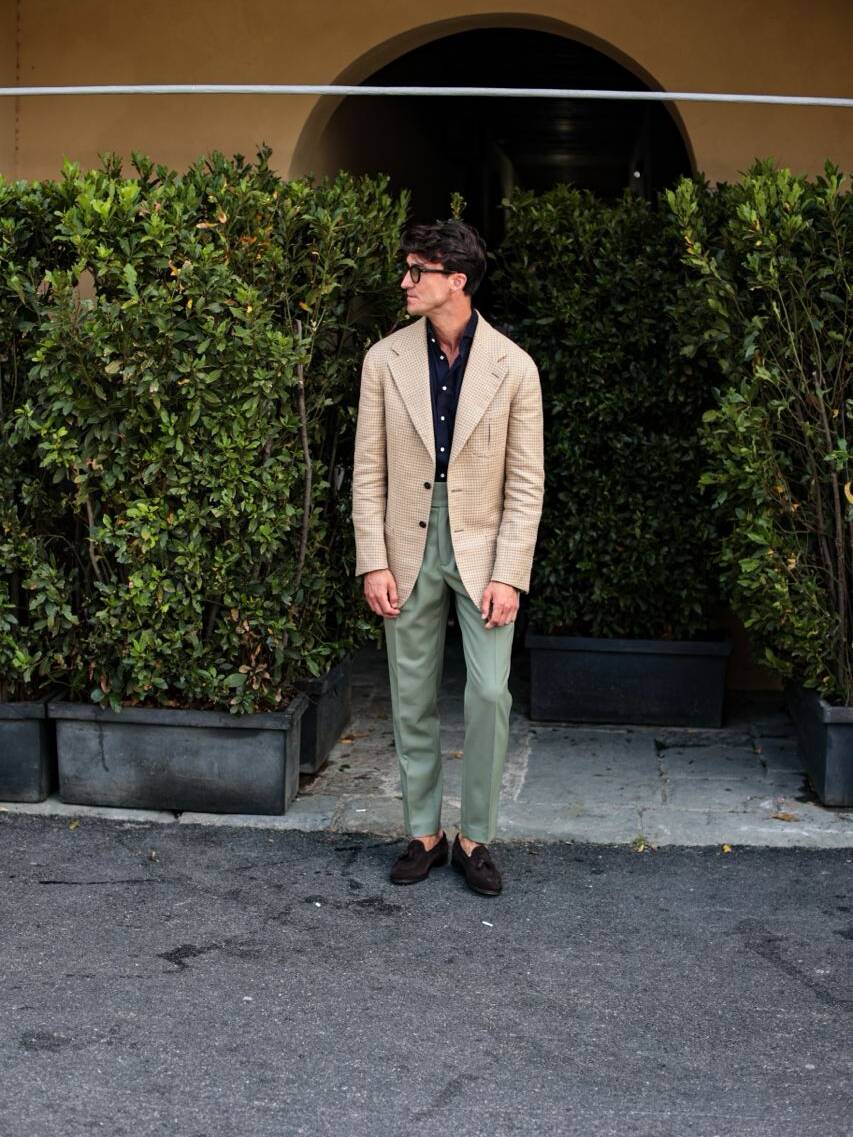 Man at Pitti Uomo summer fashion event in Italy wearing a beige bespoke jacket, dark blue shirt, and woolen olive green trousers