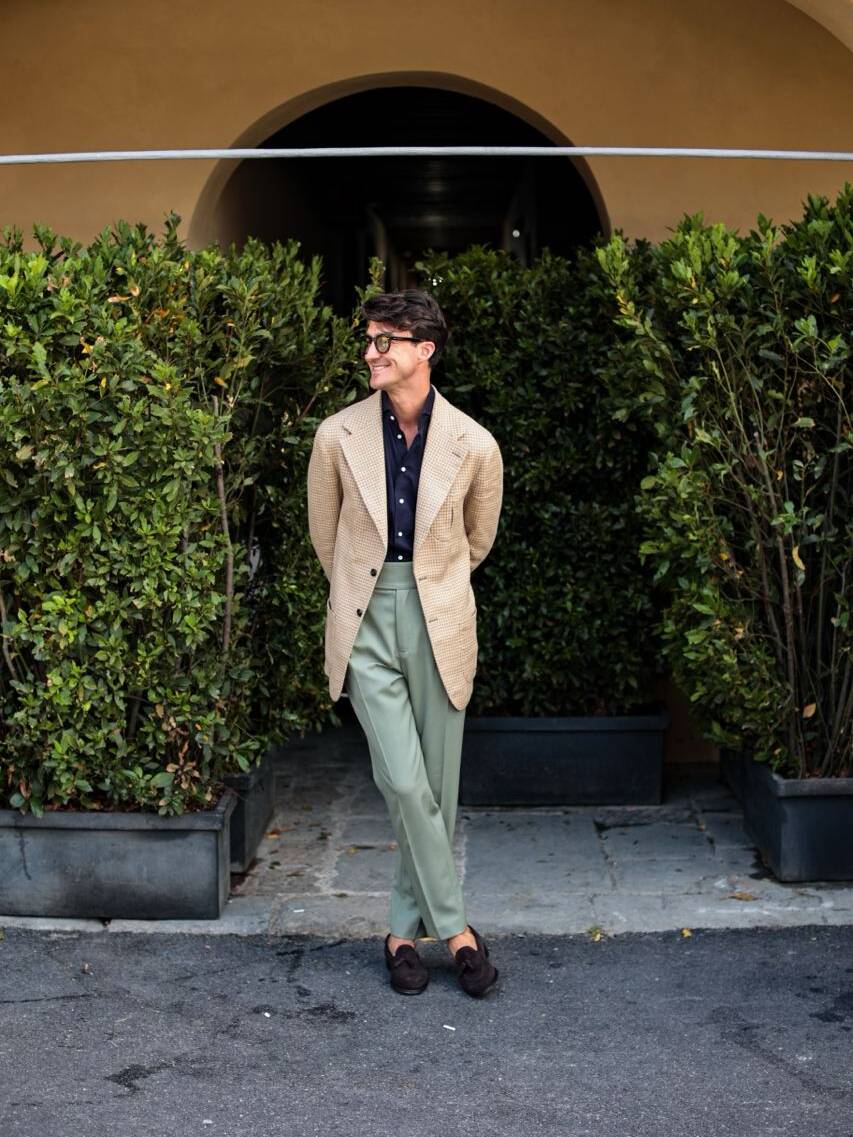 Man at Pitti Uomo summer fashion event in Italy wearing a bespoke jacket, dark blue shirt, and olive green trousers, standing in front of lush greenery