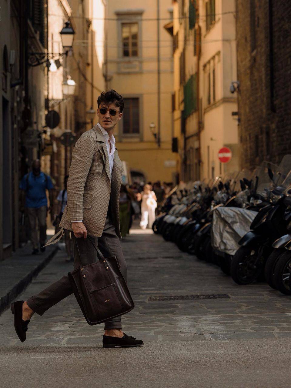 Stylish man in a beige blazer carrying a leather tote bag, walking through a European street. Leather craftsmanship and fashion