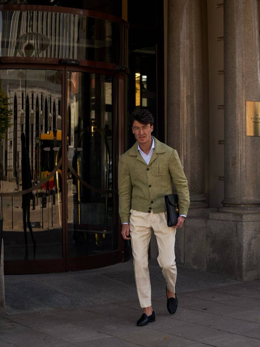 Well-dressed man in a green textured jacket and cream trousers holding a leather portfolio outside a modern building. Leathercraft elegance and gentleman style