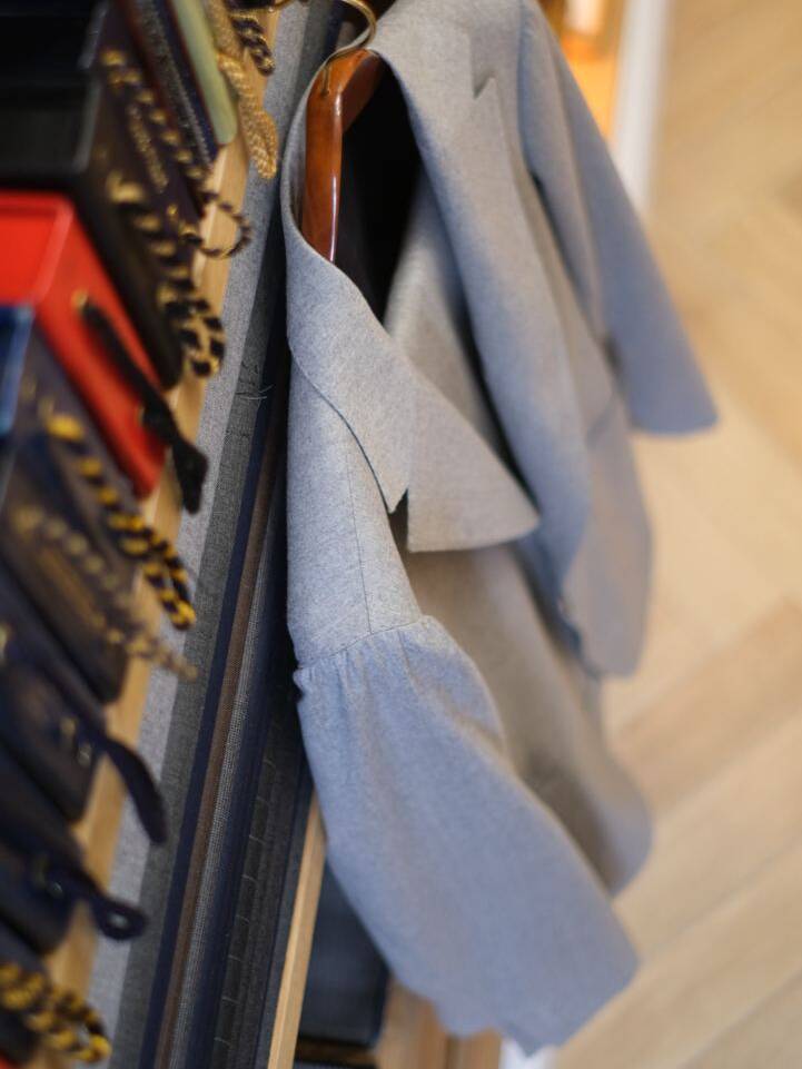 Gray suit jacket on a wooden hanger, highlighting the glued method of suit construction. Fabric swatches in the background emphasize the importance of material choice in glued suits