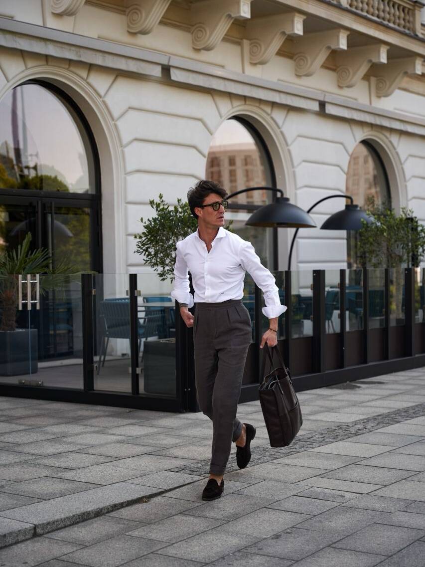 Man in tailored white shirt and dark grey double-pleated trousers walking confidently with a briefcase. The look emphasizes how to choose pants with pleats for a professional and sleek style