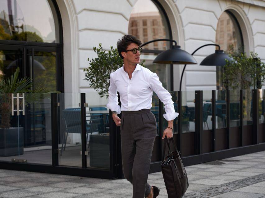 Man in tailored white shirt and dark grey double-pleated trousers walking confidently with a briefcase. The look emphasizes how to choose pants with pleats for a professional and sleek style