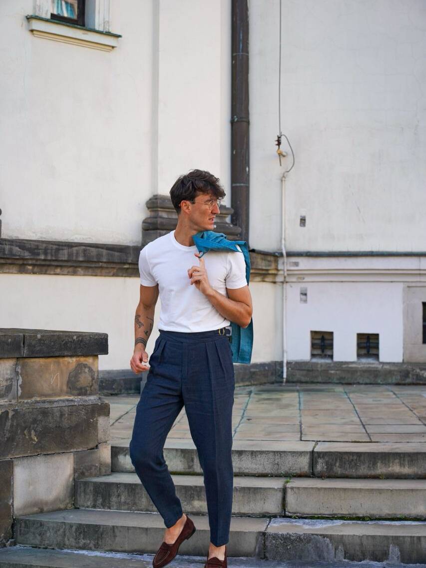 Man wearing a white T-shirt with dark blue pants with pleats, holding a blue jacket over his shoulder. A casual guide on how to choose pants with pleats for a relaxed, modern look