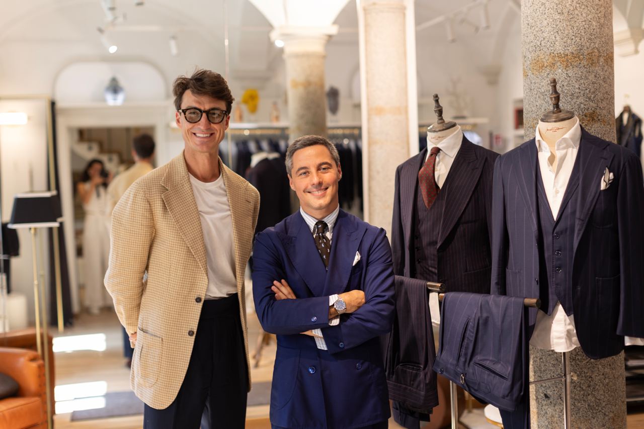 Two men pose in a stylish Milan workshop surrounded by tailored suits during a style master class. The atmosphere highlights craftsmanship, fashion, and elegance, symbolizing the essence of Italian sartorial expertise